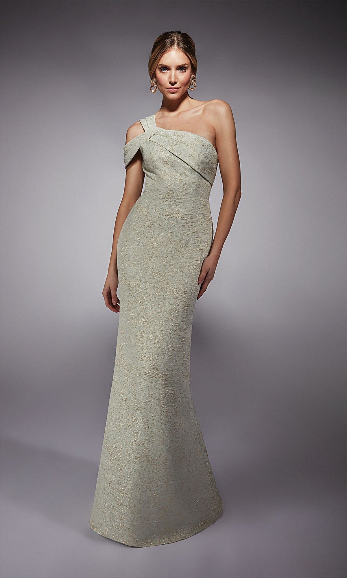 A woman models the Alyce 27749, a light-colored, floor-length formal jacquard sheath dress, standing against a plain gray background.